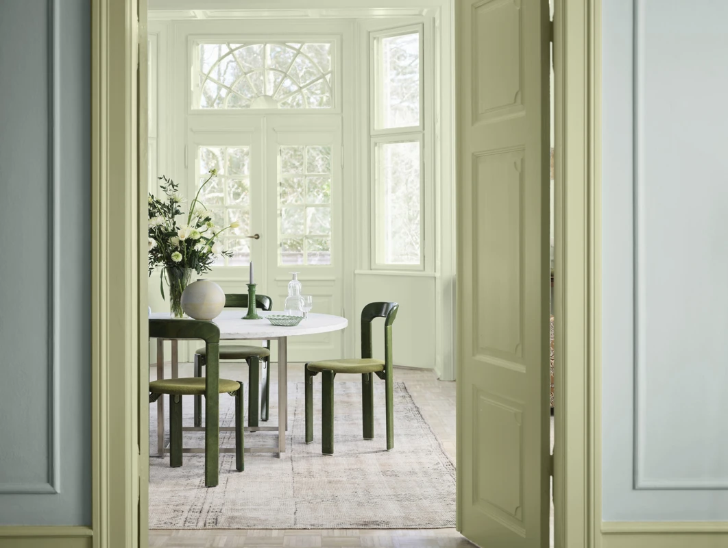 Dining room with pastel walls, a white round table, green chairs, and a vase with flowers under natural light.