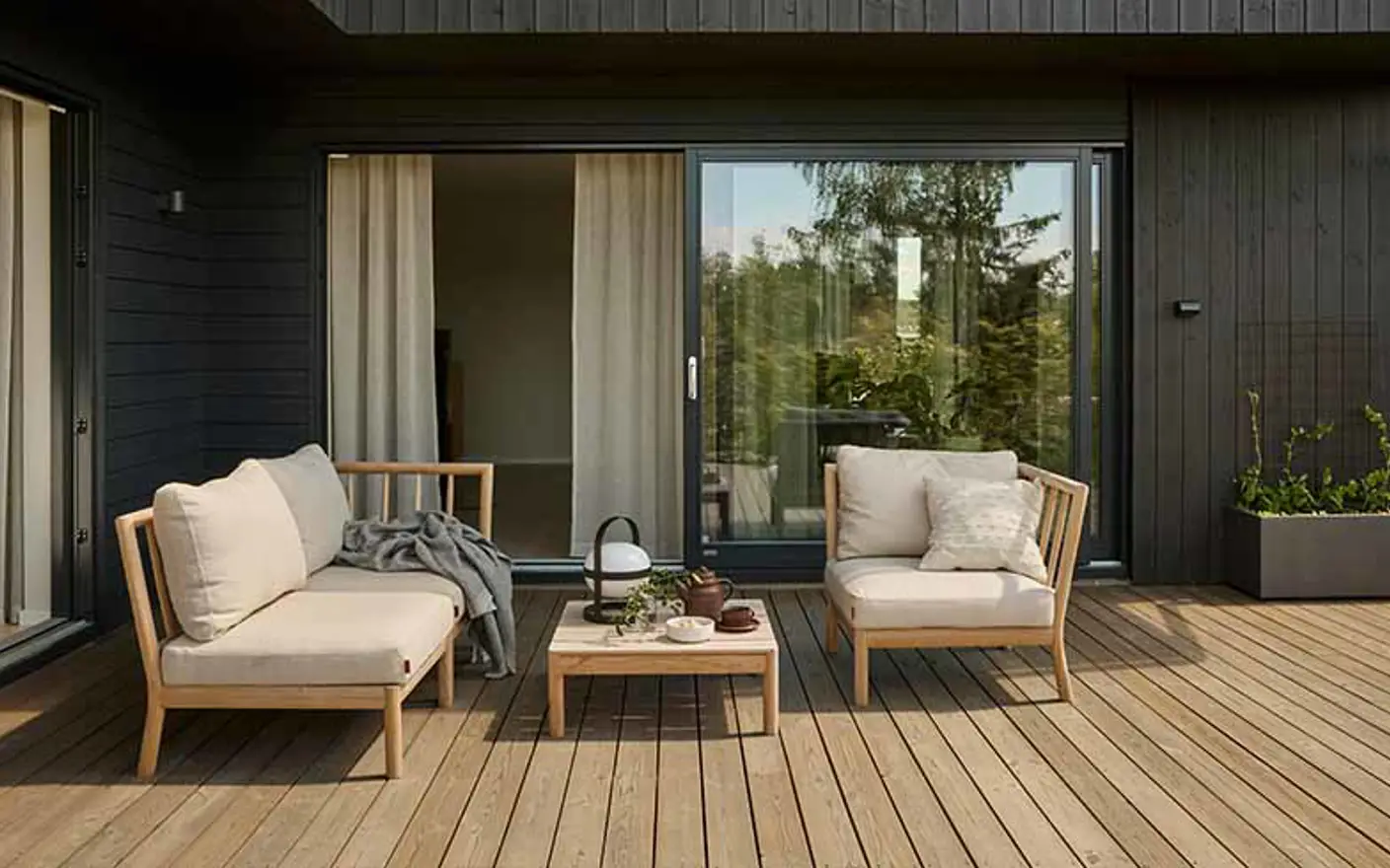 Moderne terrasse med lyst brune terrassebord i tre som er beiset med TREBITT Terrasse i fargen 9074 Nordisk Tre, to komfortable lenestoler med lys beige puter, et lite trebord med te-sett, og store glasskyvedører som fører inn til huset som er malt i Jotun 90033 Funkissort