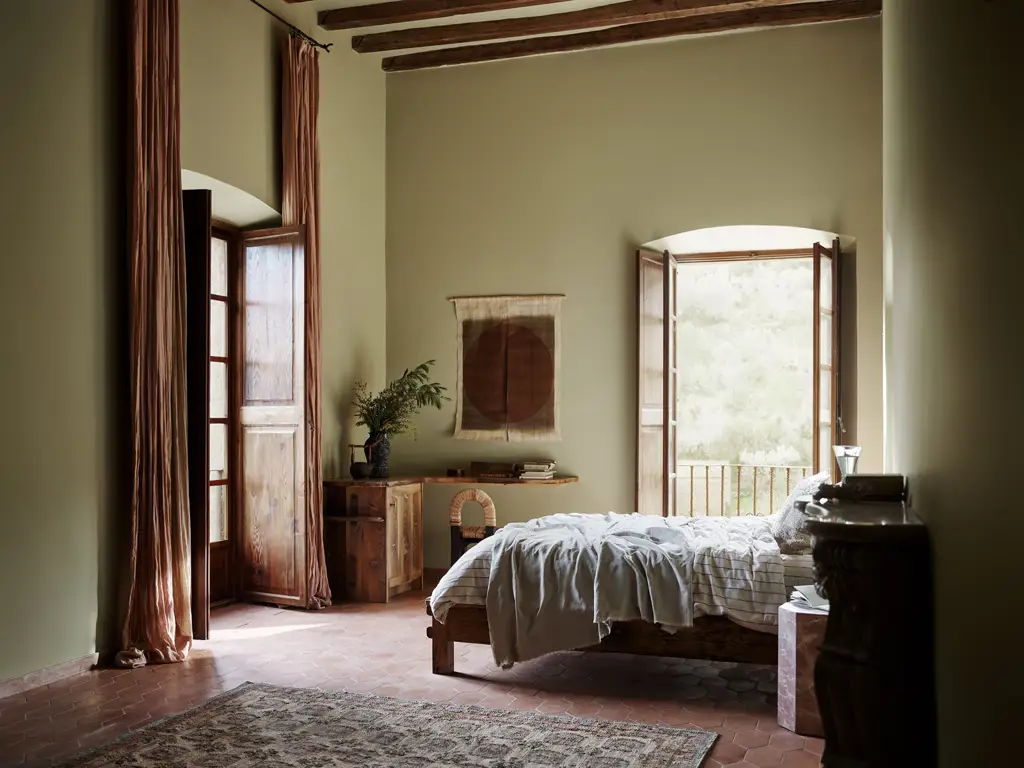 A rustic bedroom featuring earthy olive green walls in Jotun 0290 Biri, exposed wooden ceiling beams, open French doors, and a low wooden bed