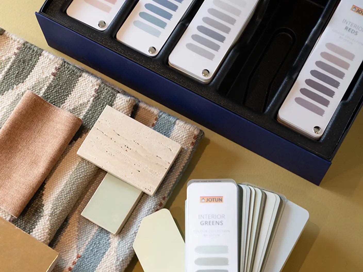 Färgsnuttar och prover från Jotun som visar alternativ för inredningsdesign, inklusive färger från Jotun i olika nyanser som blått, grönt och neutralt, visas tillsammans med tyg- och materialstrukturer på en beige yta
