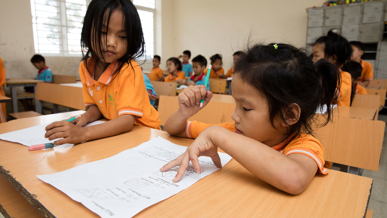 Elever i ett klassrum som arbetar med skoluppgifter. Barnen bär orangea skoluniformer och fokuserar på sina skrivuppgifter vid skrivborden