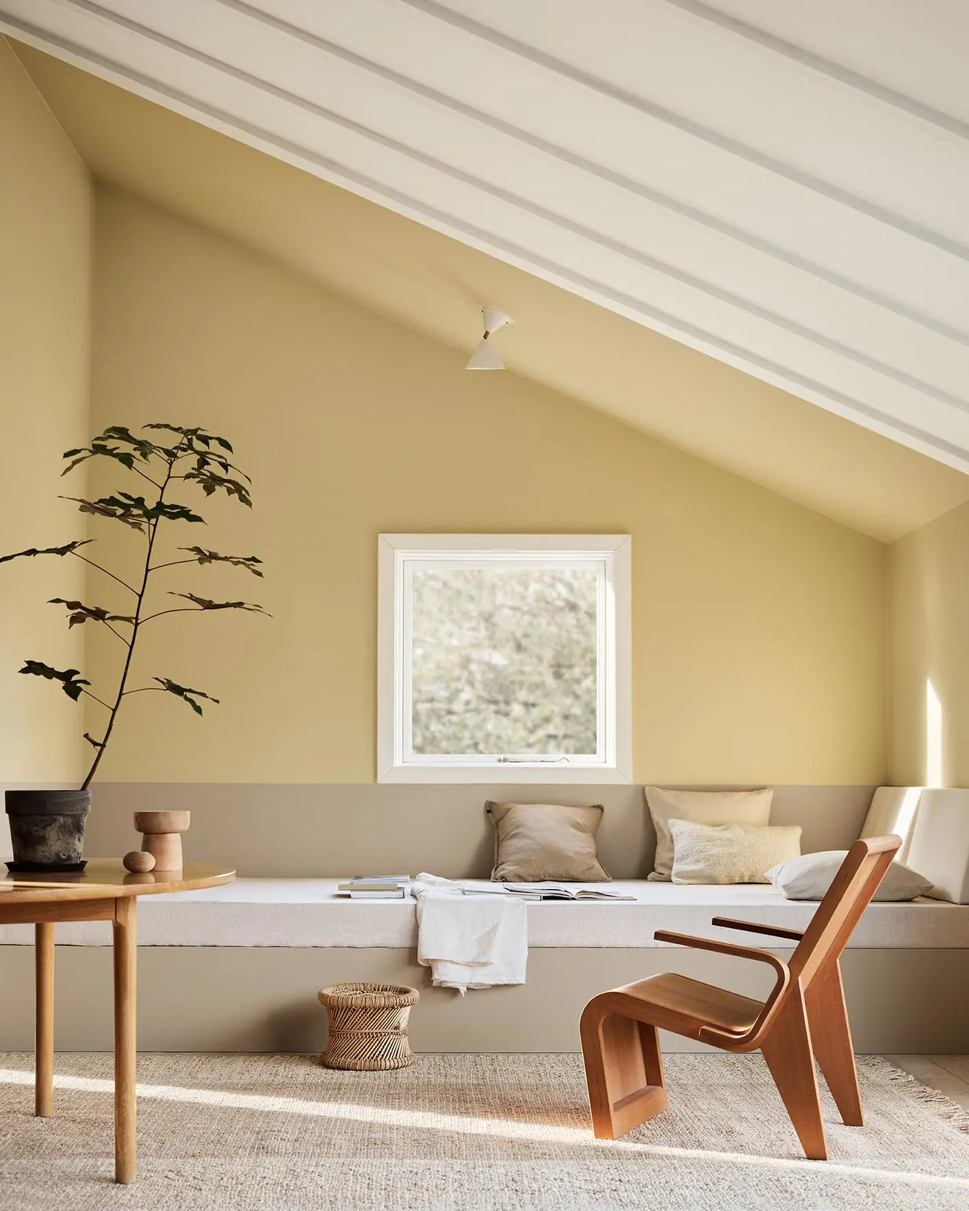 A daybed, wodden chair and a potted plant against muted, greenish yellow walls painted in the colour 1965 Ginger Tea from Jotun. Daybed painted in the colour 12300 Hazelnut Beige.