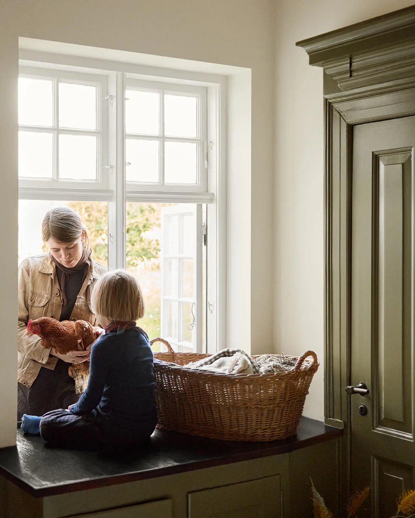 Woman and a child child on a kitchen counter by the window next to a olive-green cabinet in the colour 8597 Seaweed Green, against a muted yellow wall in the colour 12079 Gleam from Jotun.