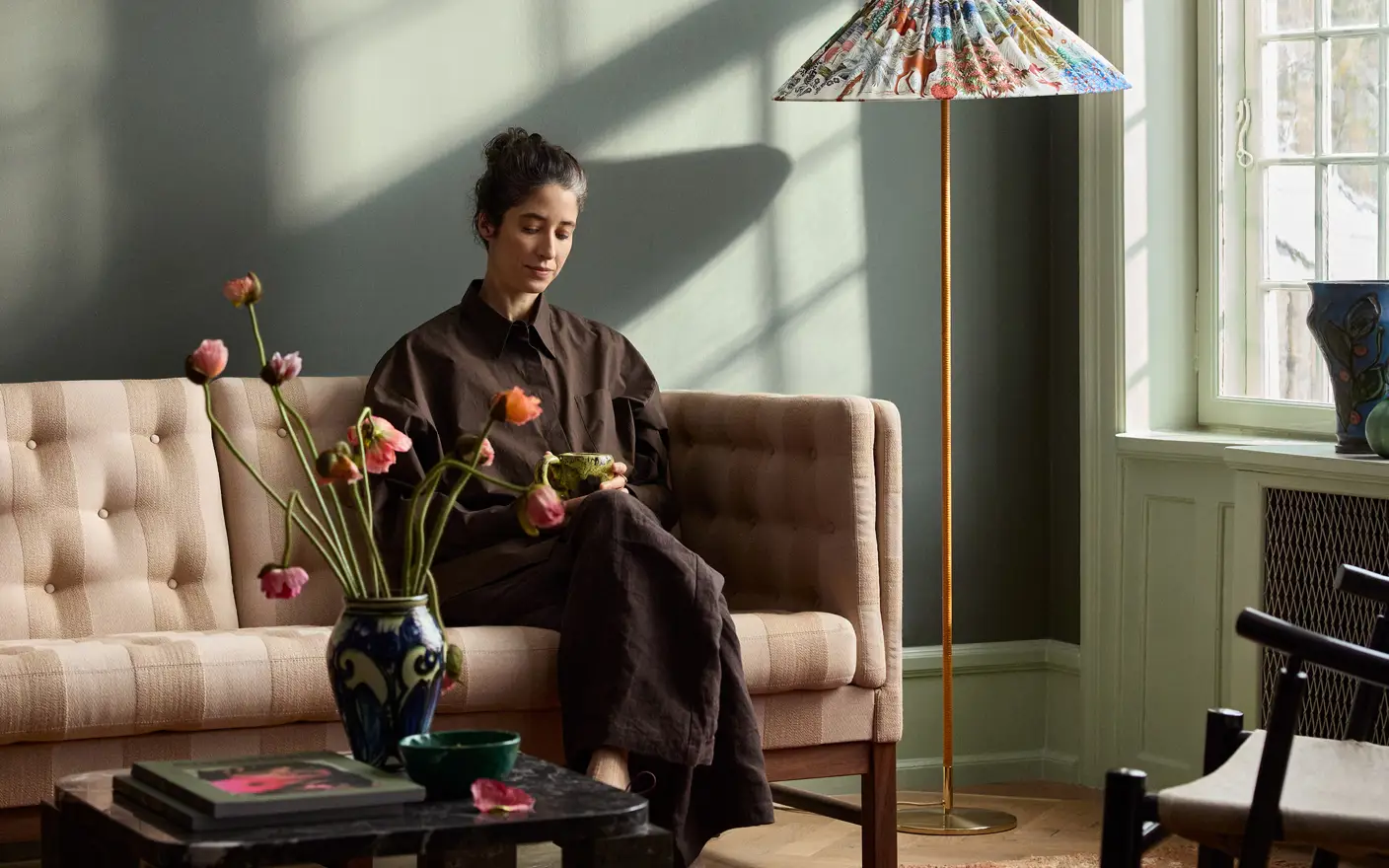 En kvinna sitter på en beige soffa, håller i en kopp, i ett solbelyst vardagsrum med en färgglad golvlampa och en blomvas på ett soffbord. Vägg målad i Jotun 7613 Northern Mystic.