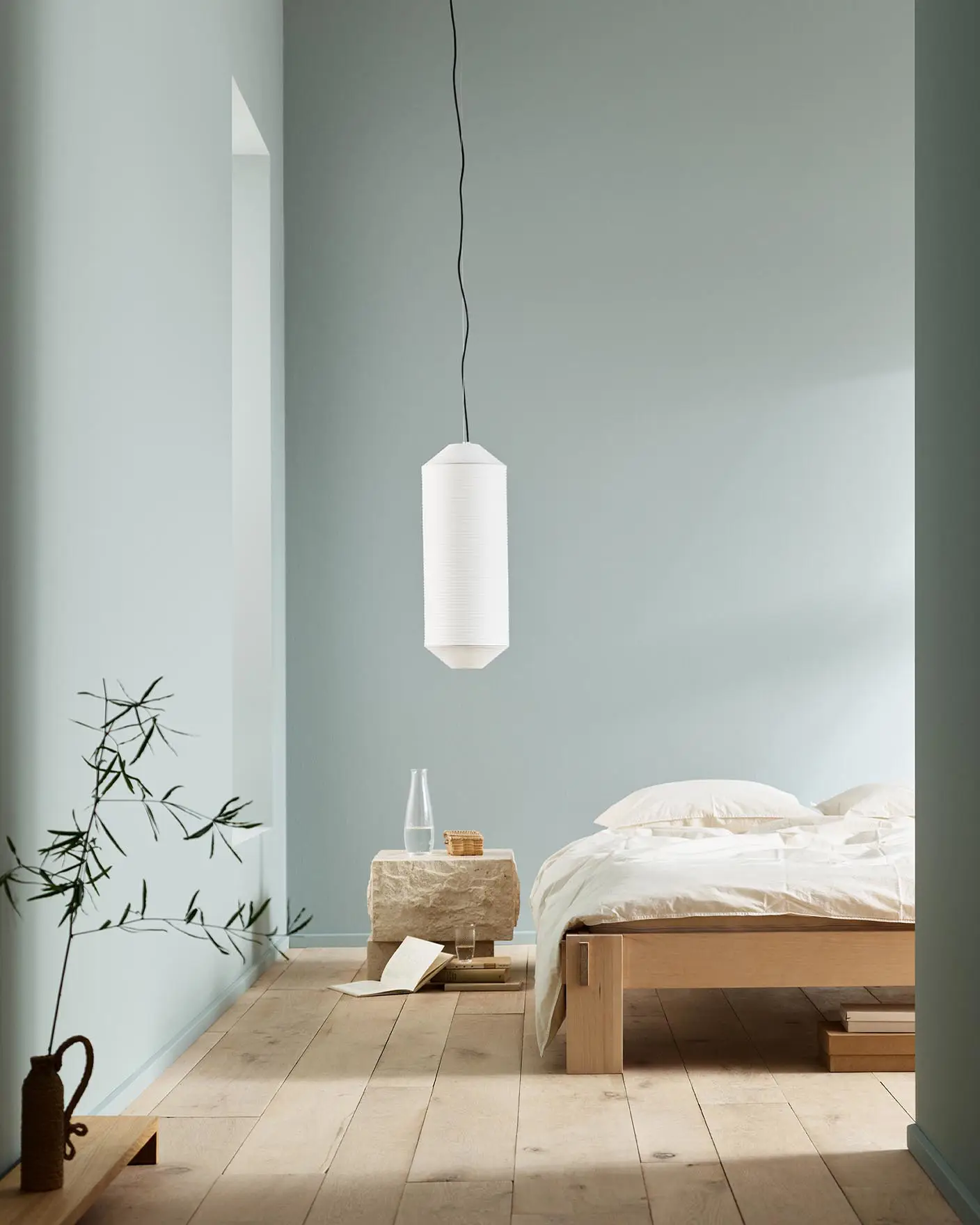 Minimalist bedroom with white bedding, a stone side table, and a plant against muted blue walls painted with the colour 6378 Iconic from Jotun.