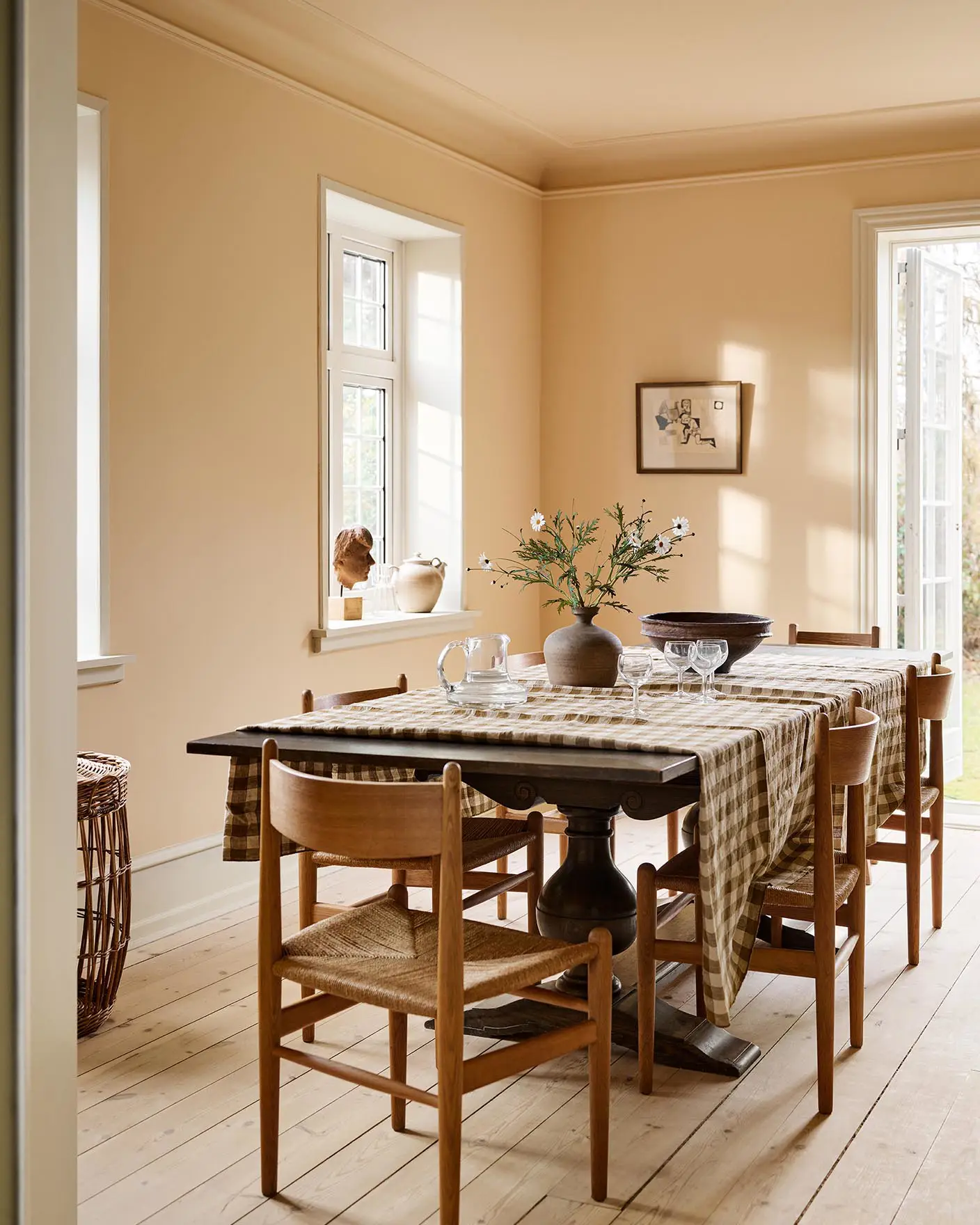 Dining room with a checkered tablecloth, wooden chairs, against an ochre yellow wall painted in the colour 11202 Mild Ochre from Jotun.
