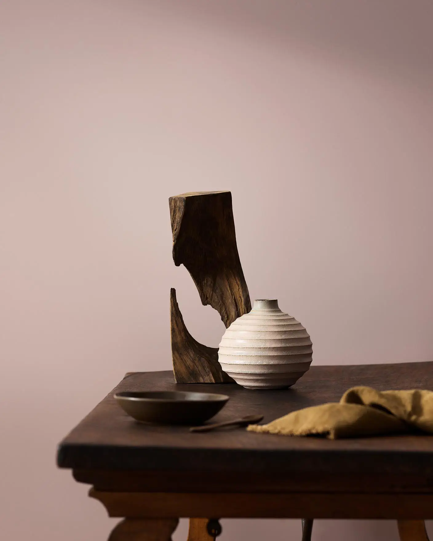 Wooden table with a ceramic vase, sculpted wood, and a dark bowl against a muted pink background painted with the colour 20054 Silky Pink from Jotun.