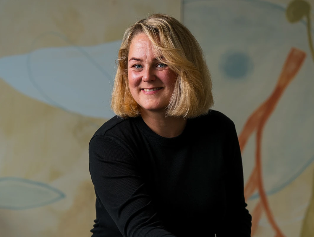 Maria Horgen, regional fargesjef i Jotun, smilende foran en dekorert vegg i ulike farger, med fargeprøver foran seg på et bord