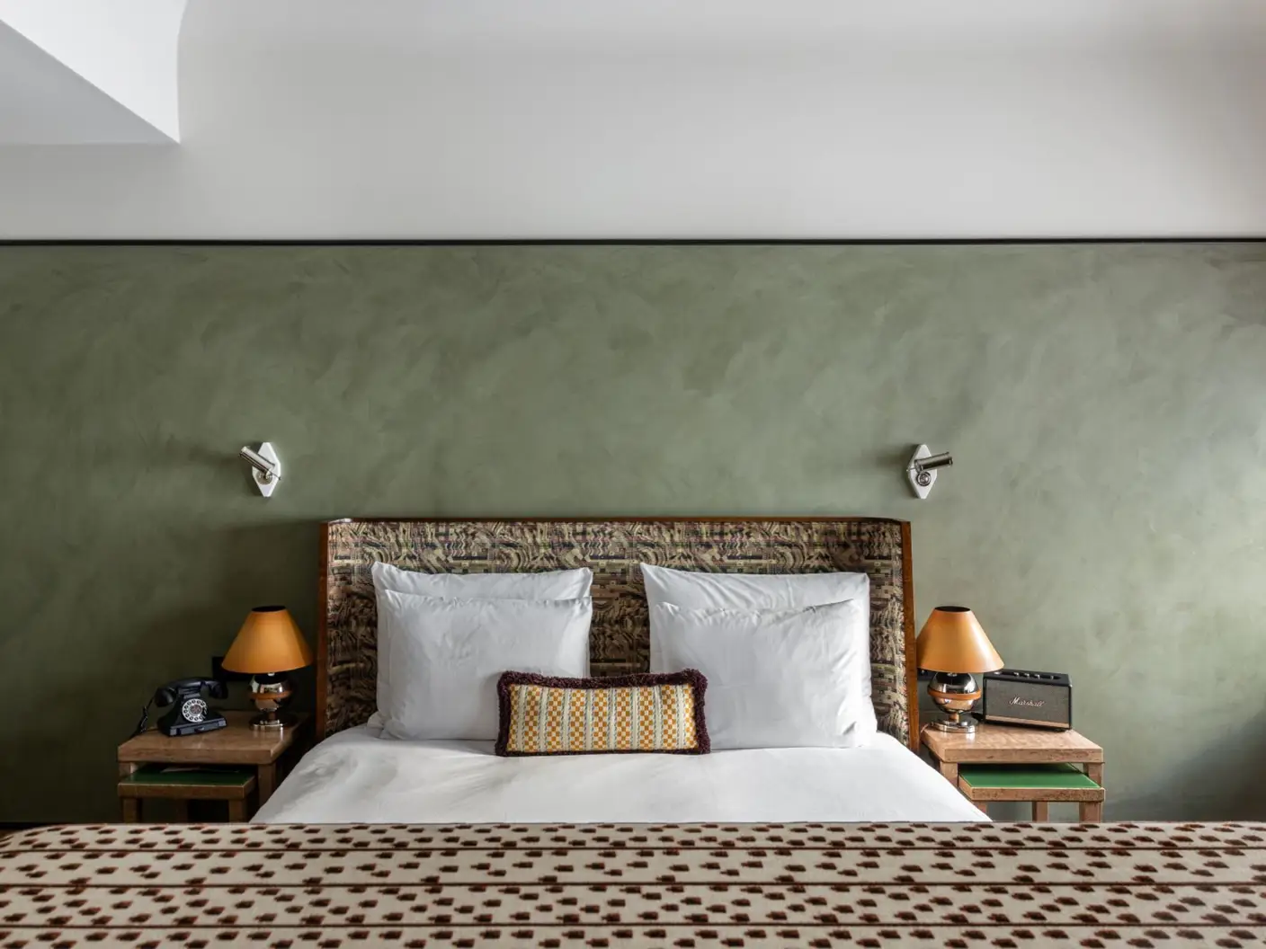 Koselig hotellrominteriør med en rustikk sengegavl, hvitt sengetøy, dekorative puter, grønn kalkmalt vegg med LADY Minerals kalkmaling i fargen , vintage telefon, bordlamper og radio på nattbord