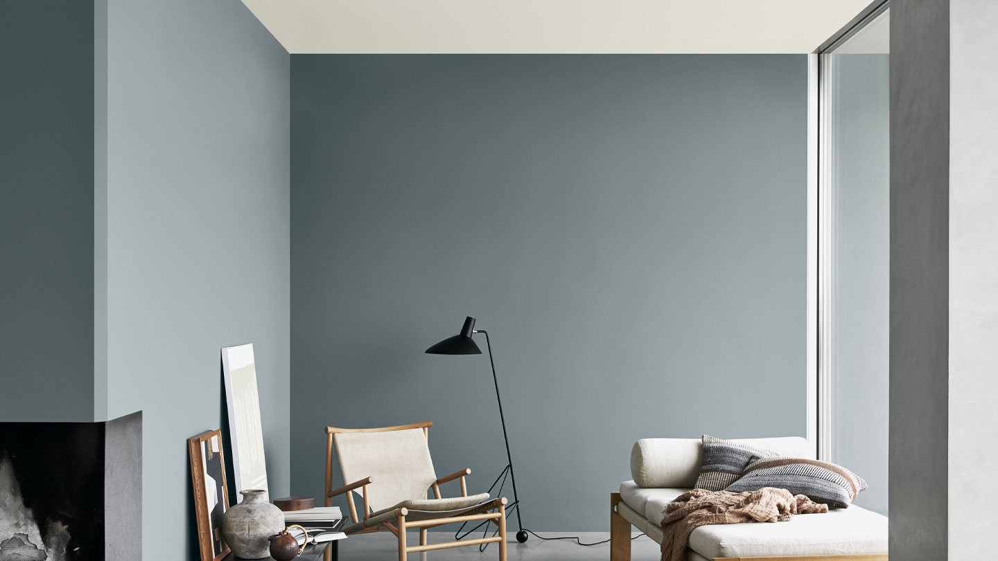 Moderne stue med grå vegger, minimalistiske møbler, inkludert en lenestol, svart lampe og beige sofa med strikkede puter, store vinduer som slipper inn naturlig lys