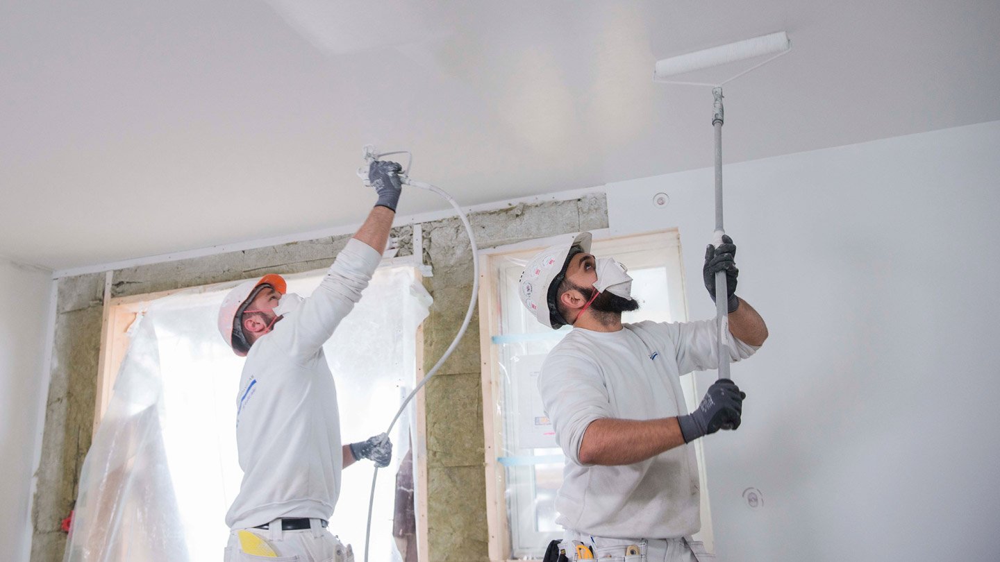 To håndverkere maler et rom, den ene med malingssprøyte og den andre med malerulle. Begge bruker beskyttelsesutstyr og jobber på et tak. Renovasjon og innredning