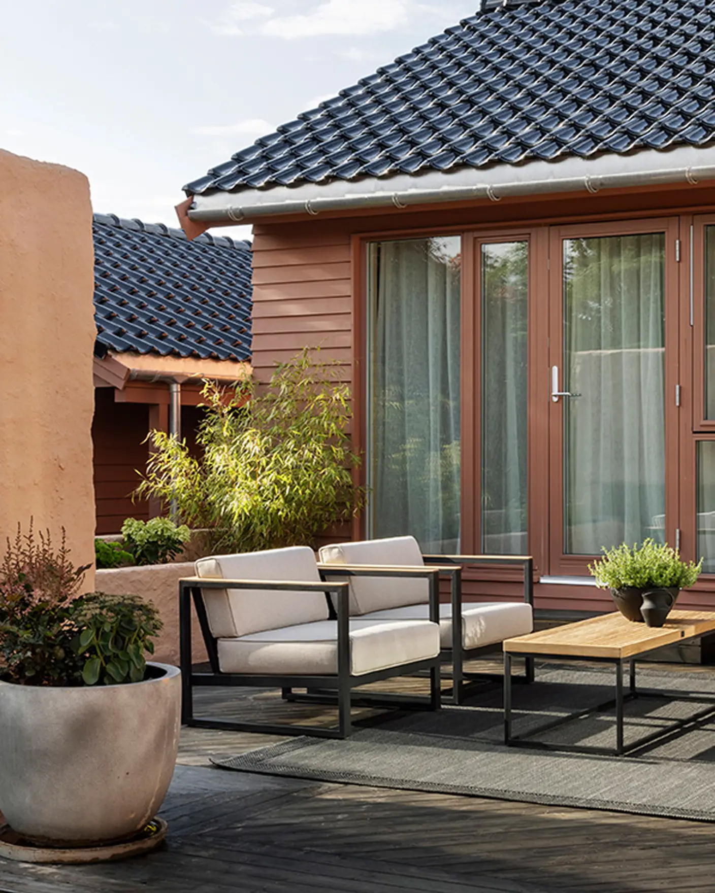 Moderne terrasse beiset med TREBITT Terrassebeis, med stilfulle hagemøbler og frodige planter foran et rødrosa hus malt i Jotun Amber Red med mørkt tak og store vinduer