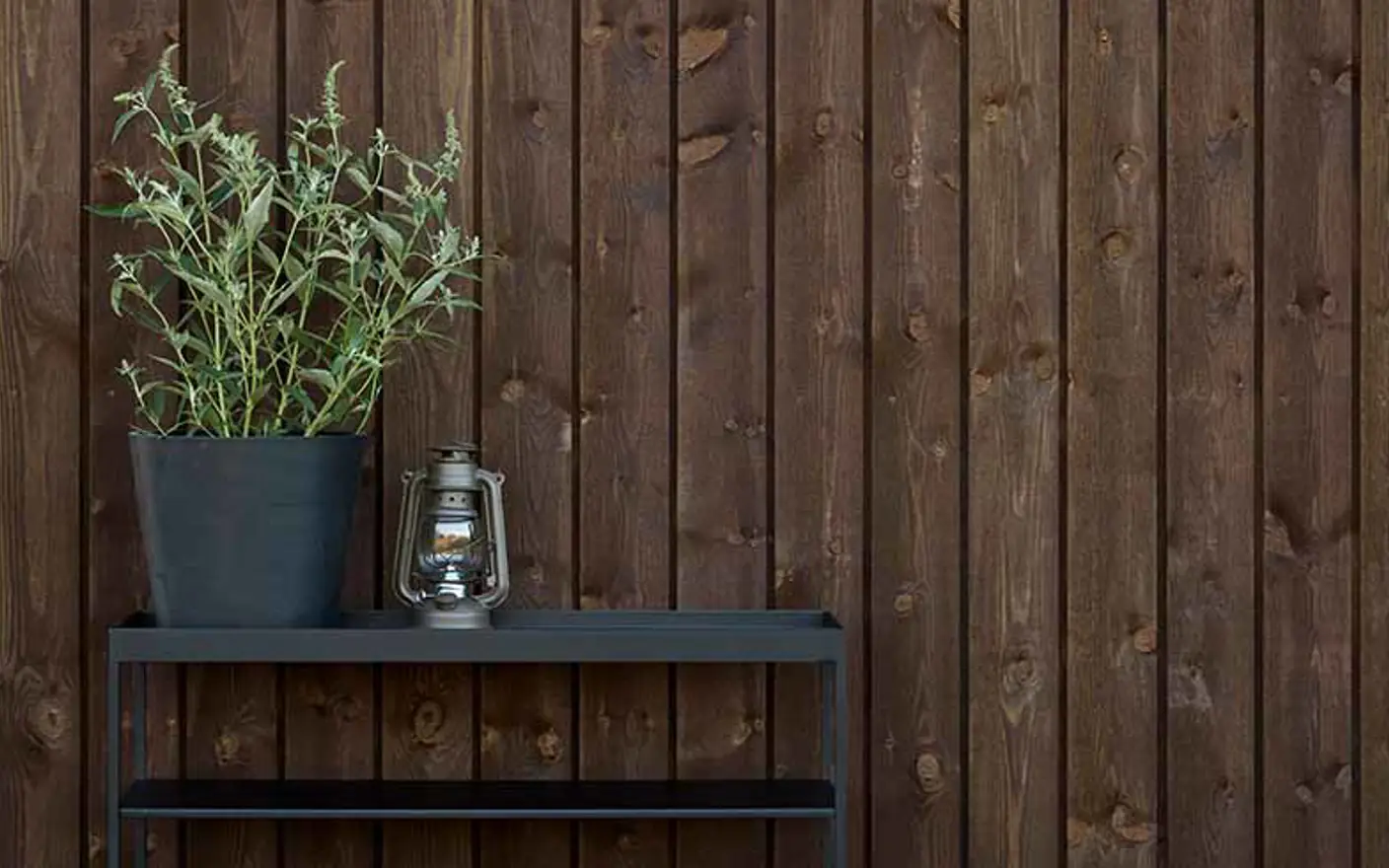 Planter i svart potte og lykt på et moderne, svart metallbord foran en mørk trevegg beiset i TREBITT 9155 Rich Brown