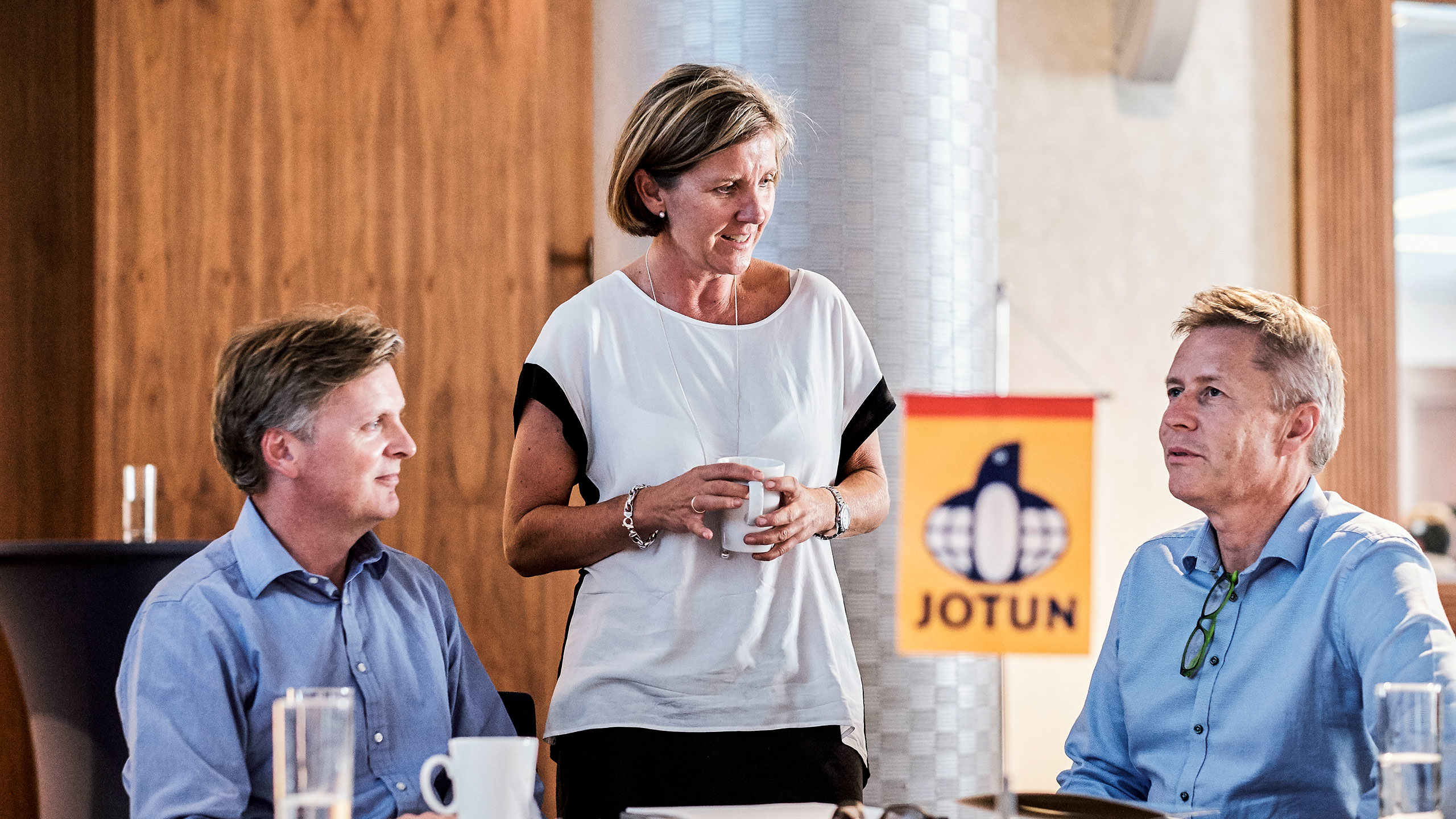 Tre personer i ett möte vid ett bord med Jotun-logotypen i förgrunden. En kvinna står och håller en kopp medan två män sitter ner och lyssnar