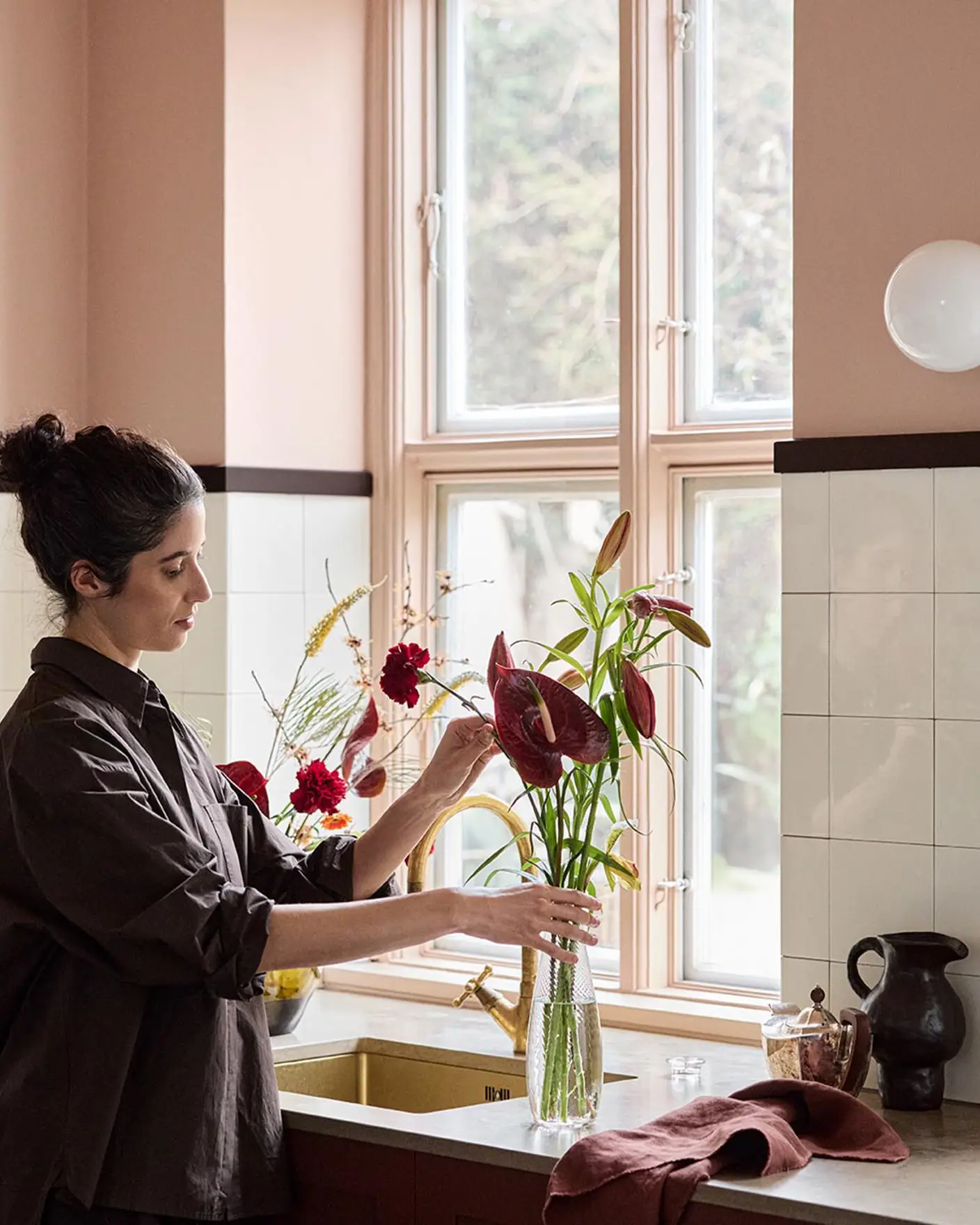 Woman arranging flowers on a kitchen counter with soft pink walls painted with the Jotun colour 20362 Pink Ambience and white tile backsplash.